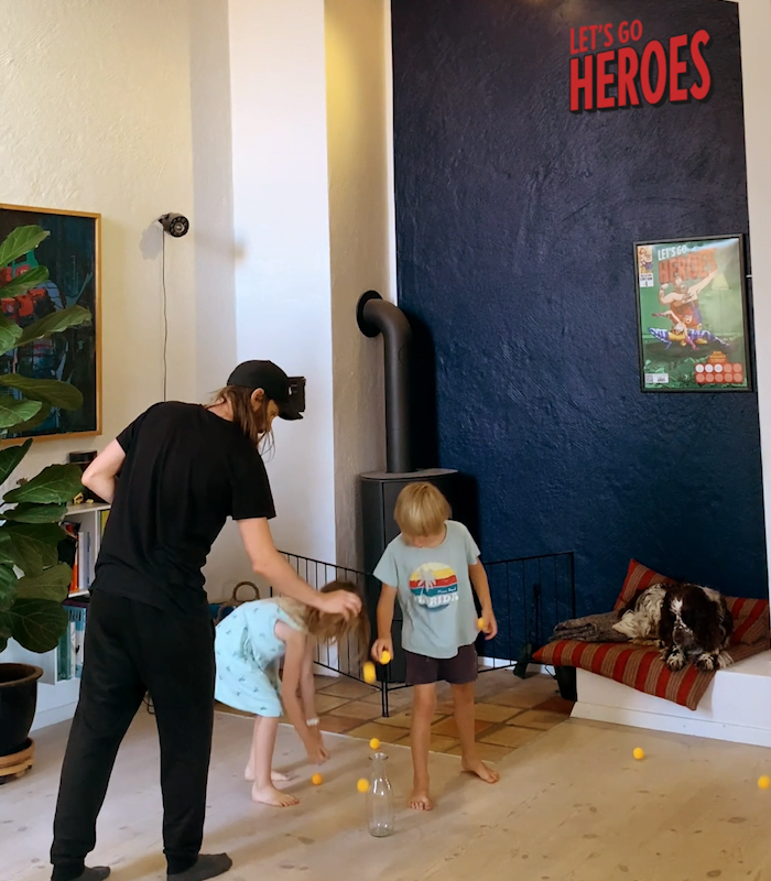 En voksen og to børn dropper bordtennis bolde ned i vase som står på gulvet. 